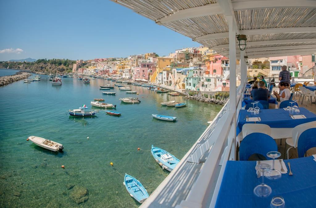 Terrace dining area overlooking the colorful waterfront village of Procida with moored fishing boats in the crystal-clear turquoise bay.