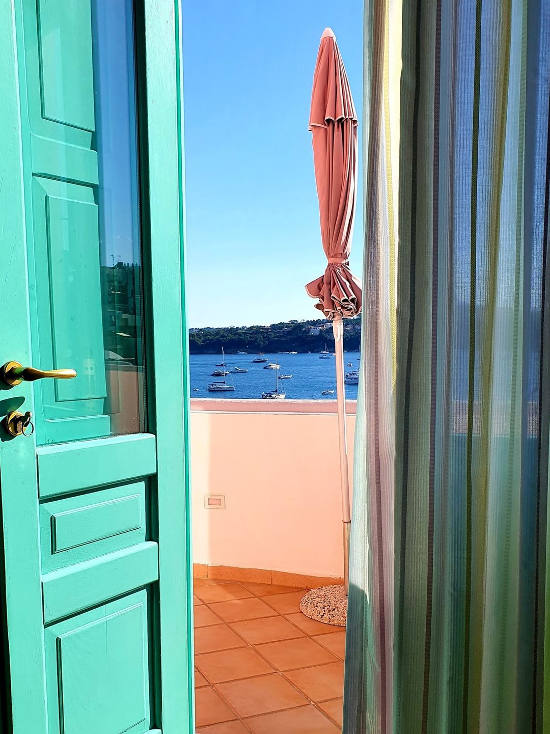 A vibrant turquoise doorway frames a coastal terrace overlooking a bay with moored boats and a closed coral-red umbrella.
