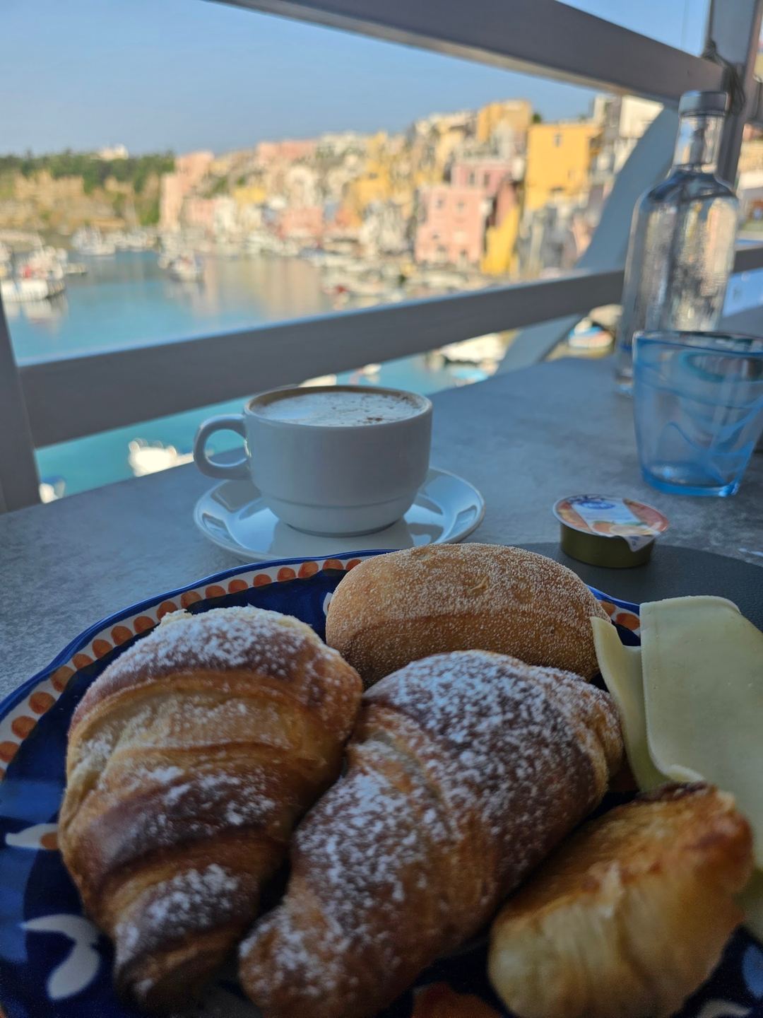 Breakfast pastries and coffee served on a terrace overlooking a colorful Mediterranean coastal village with turquoise waters.