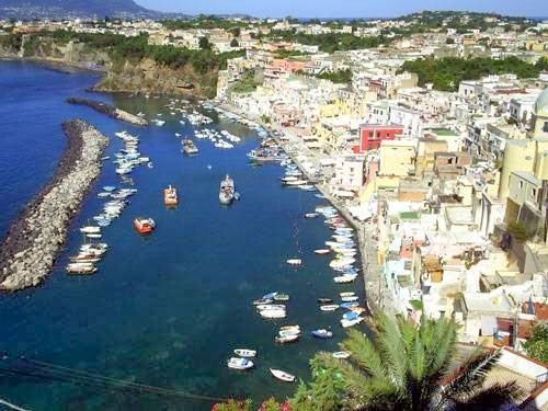 Aerial view of Corricella harbor in Procida, Italy, showing traditional colorful buildings cascading down the cliffs with numerous boats anchored in the crystal blue Mediterranean waters.