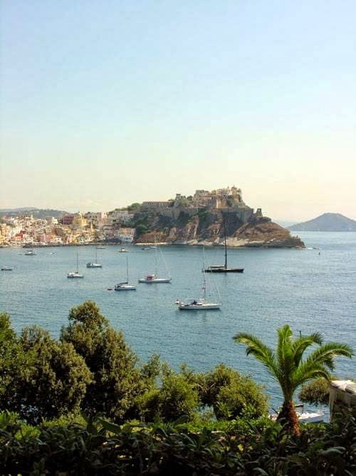 A scenic coastal view of a Mediterranean harbor with sailboats anchored in turquoise waters, a fortified island with historic architecture, and a charming waterfront village nestled along the shoreline.