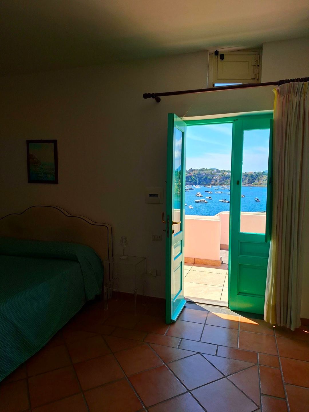 A bright bedroom with green doors opening onto a terrace overlooking a Mediterranean bay with moored boats.